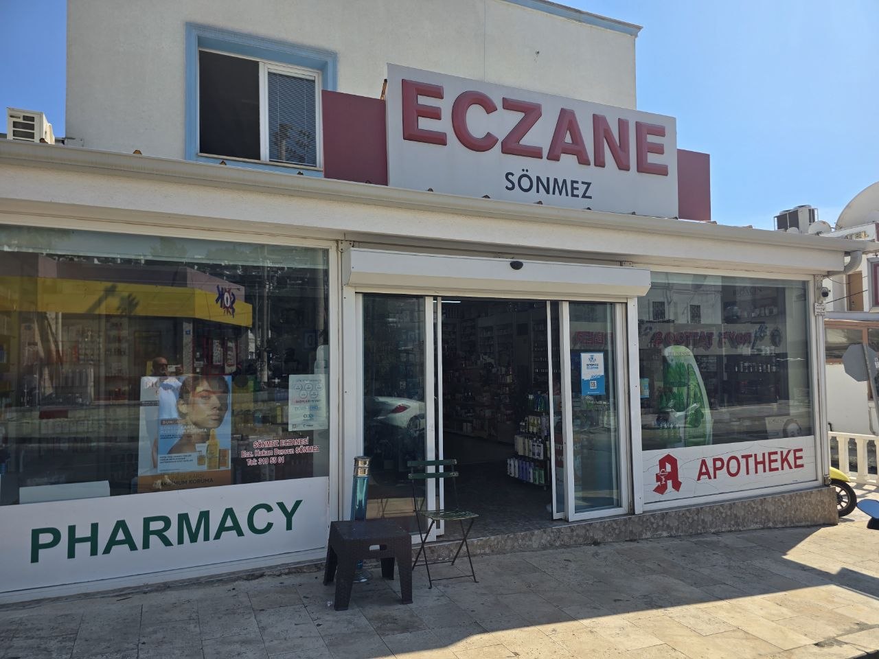 Sönmez Eczanesi, Gümbet: beyaz binada kırmızı ECZANE SÖNMEZ tabelası, camlarda PHARMACY ve APOTHEKE yazıları